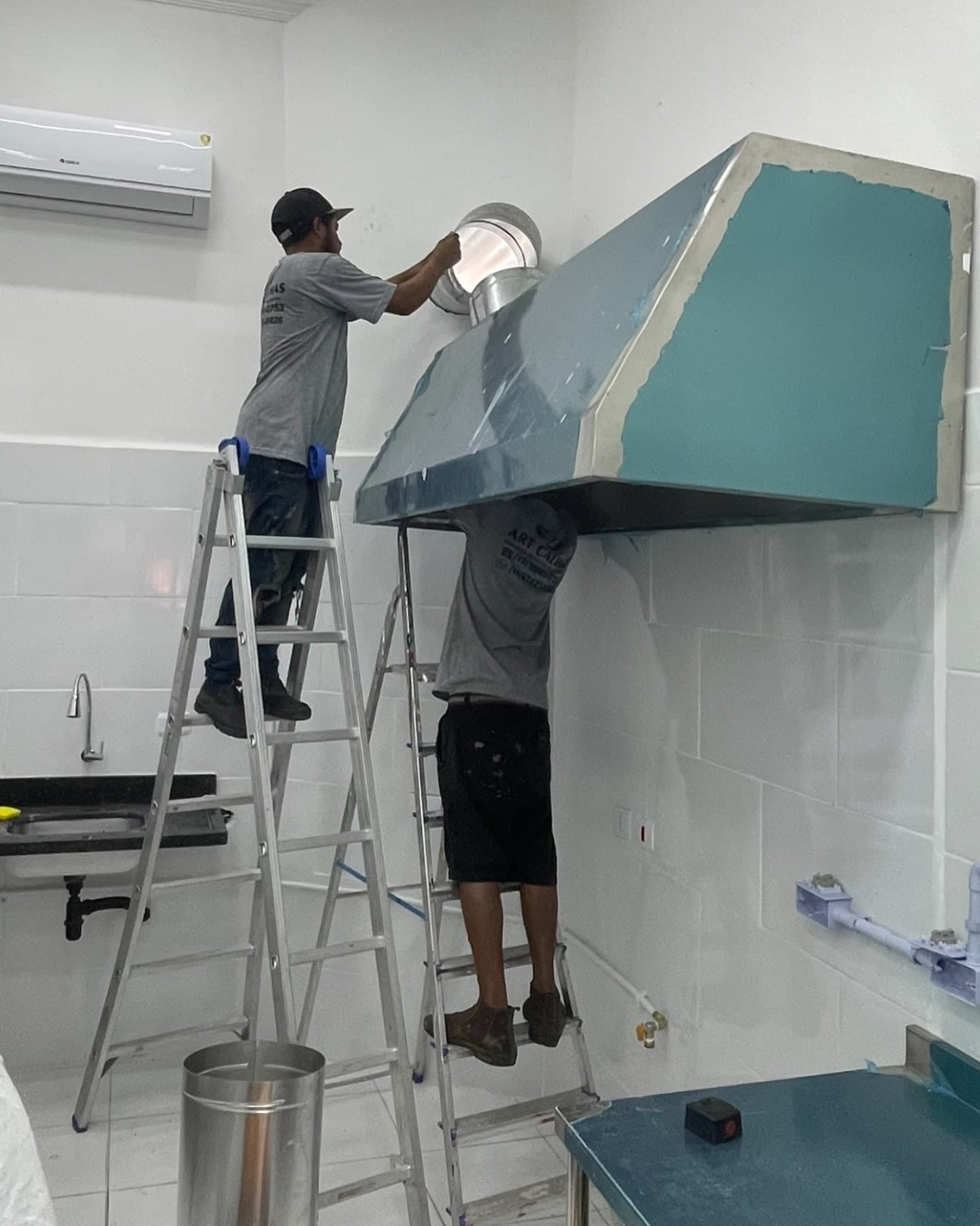Dois instaladores montando uma coifa em aço sobre a bancada de uma cozinha comercial